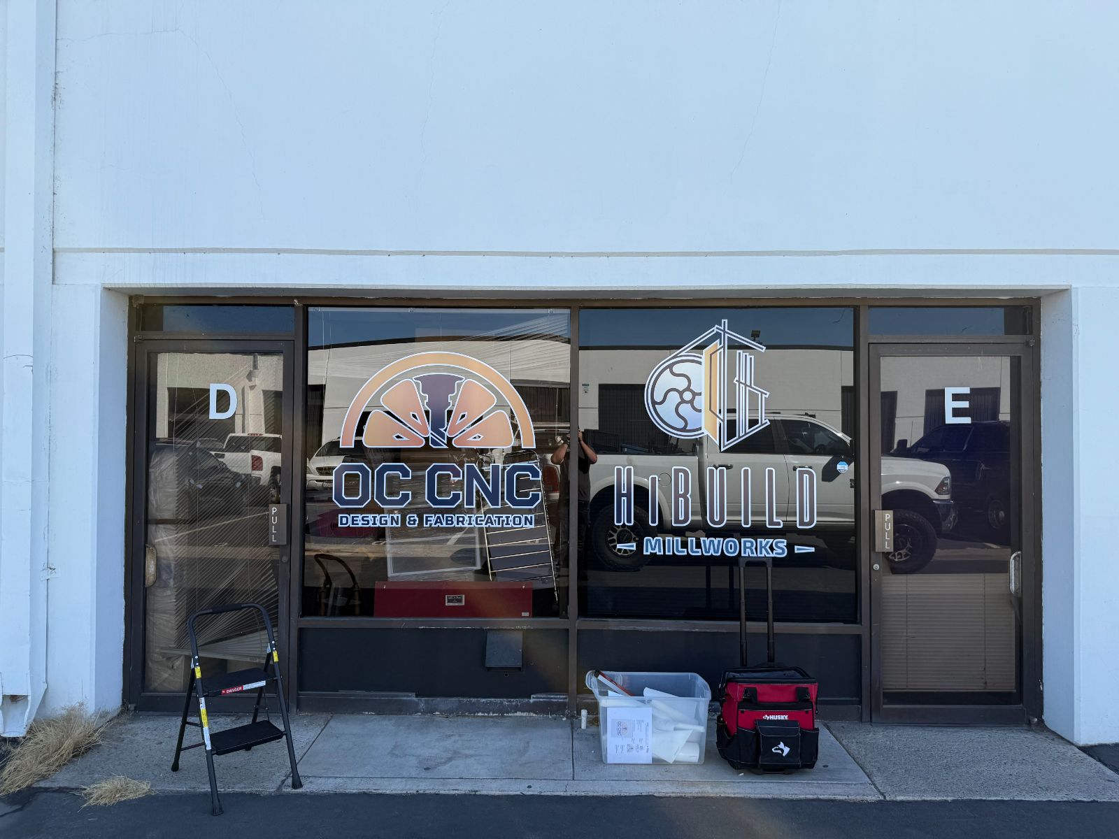 Industrial unit storefront glass with two tenants' printed vinyl logos applied to the windows: 'OC CNC Design & Fabrication' and 'HiBuild Millworks', beside doors labeled D and E, installed by Hi Sign, Brea, CA