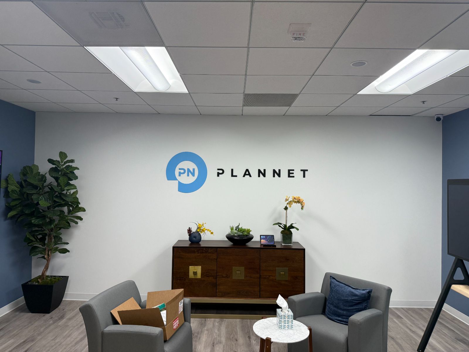 Reception wall in an office lobby with the PLANNET logo: a blue speech-bubble 'PN' mark beside black flat-cut letters spelling PLANNET, above a wood credenza, installed by Hi Sign, Brea, CA