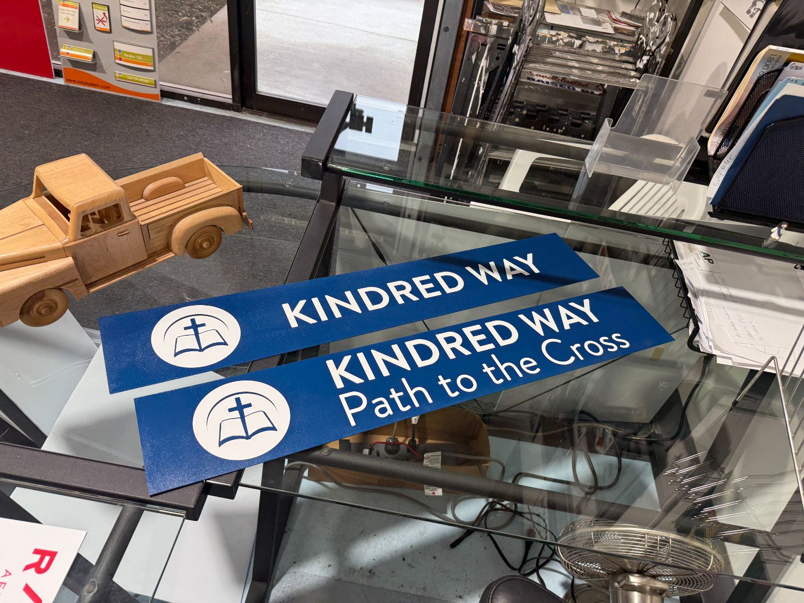 Two blue directional signs on a shop counter reading 'Kindred Way' and 'Kindred Way: Path to the Cross,' each with a white circular open-book-and-cross emblem, printed on aluminum blanks, made by Hi Sign, Brea, CA