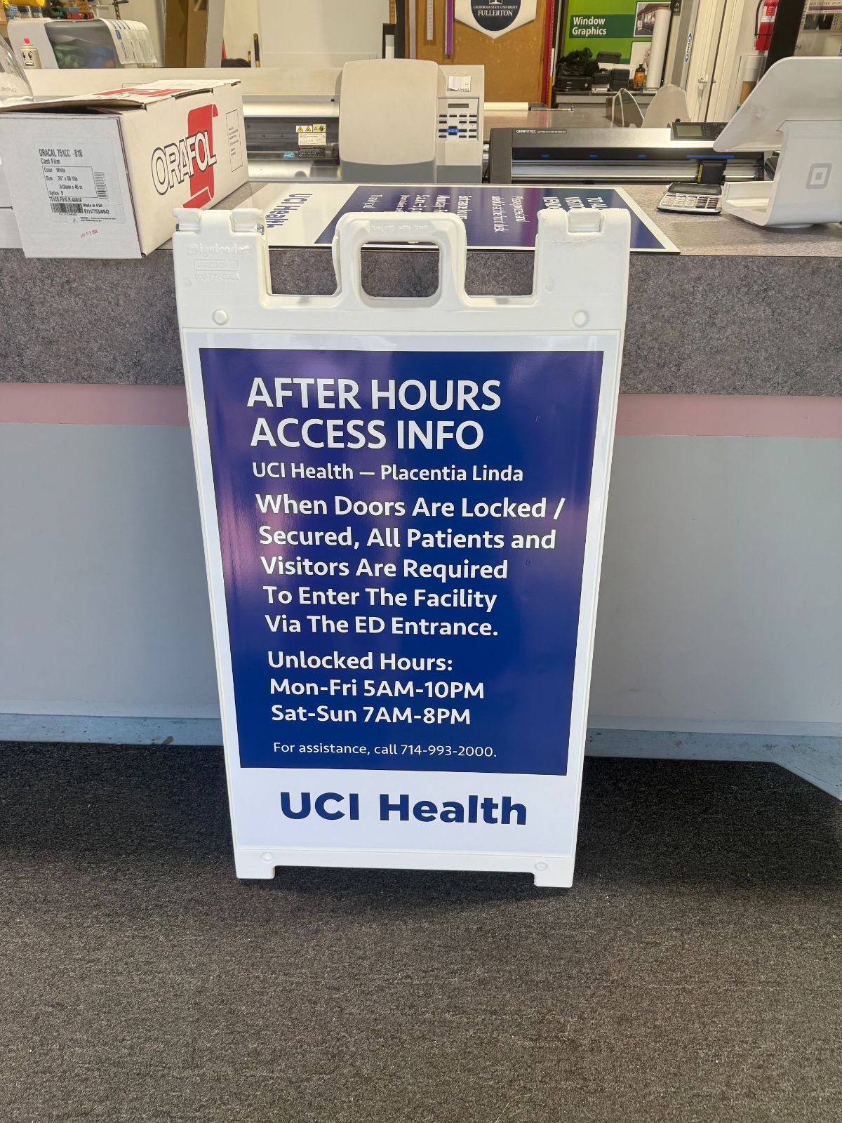White plastic A-frame sidewalk sign with a blue printed 'UCI Health: After Hours Access Info' panel listing entry instructions and unlocked hours, standing at the shop counter, made by Hi Sign, Brea, CA