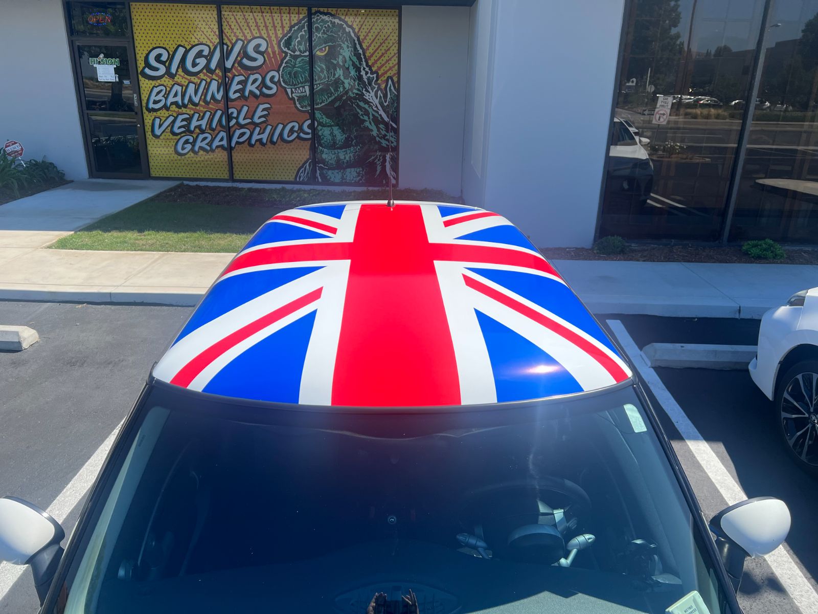 Roof of a MINI Cooper wrapped in a printed Union Jack flag graphic, parked in front of the shop window that reads 'Signs, Banners, Vehicle Graphics', installed by Hi Sign, Brea, CA