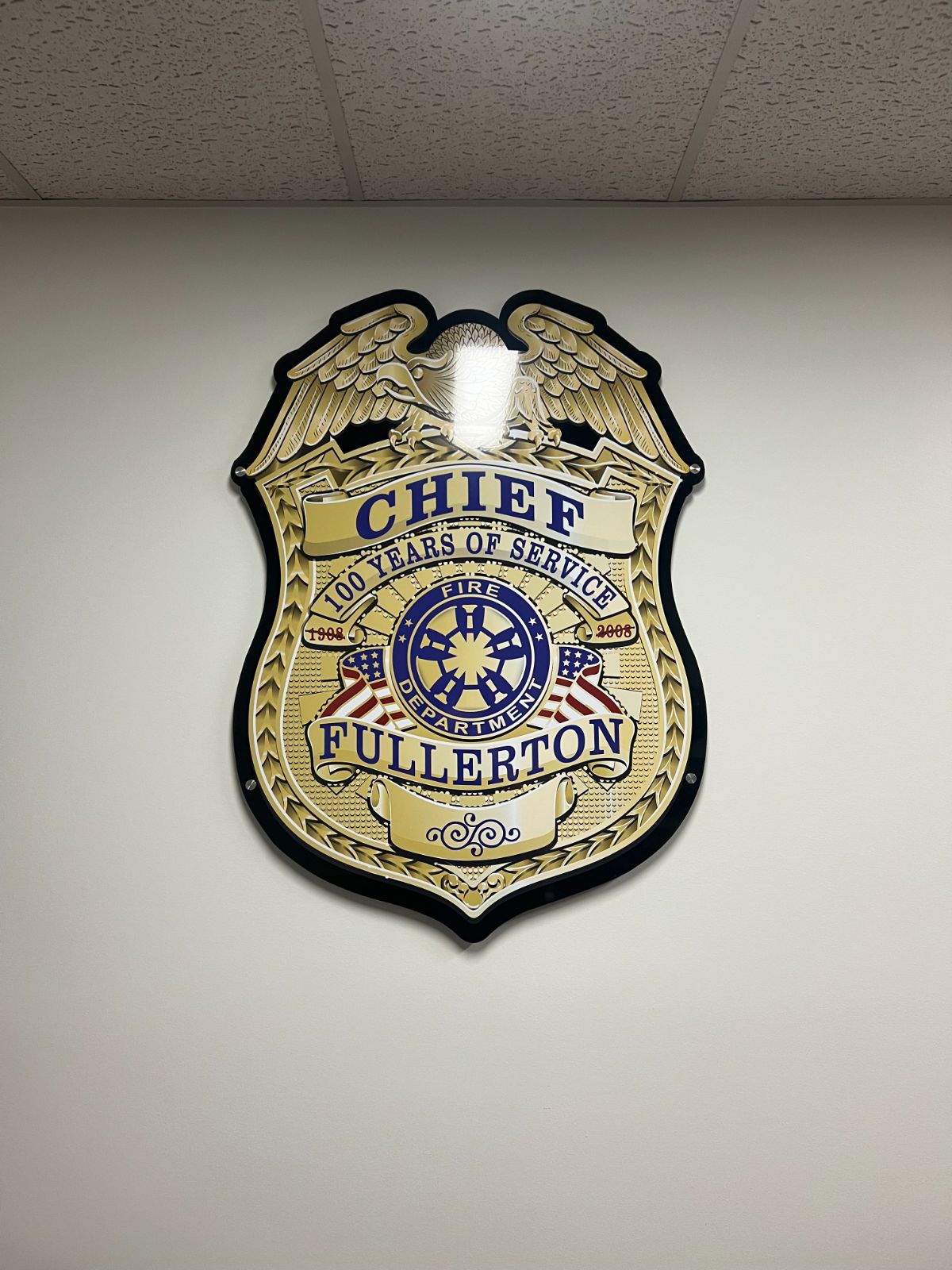 Large badge-shaped wall plaque reading 'Chief · Fullerton · Fire Department · 100 Years of Service': a full-color fire chief badge printed on clear acrylic and standoff-mounted on an interior wall, installed by Hi Sign, Brea, CA