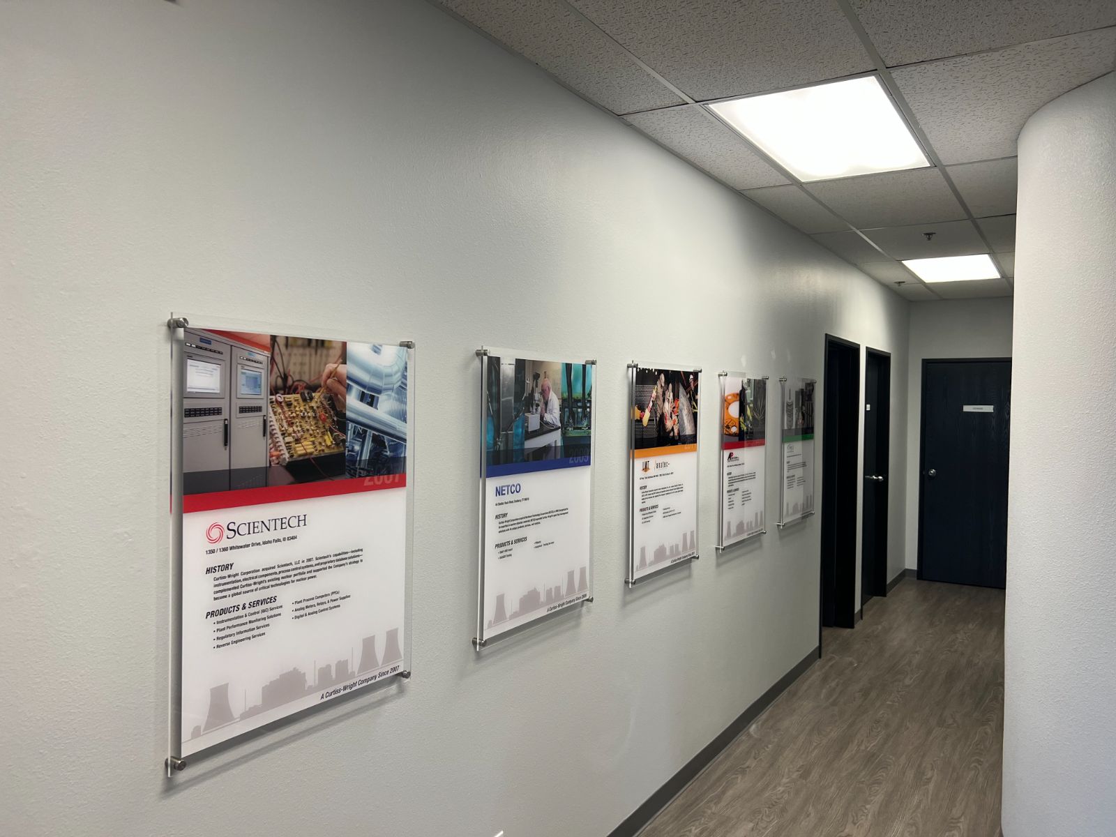 Corporate history wall in an office corridor: a row of printed company milestone panels (Scientech, NETCO, and other Curtiss-Wright brands) mounted in clear acrylic standoff frames along a white interior hallway, installed by Hi Sign, Brea, CA