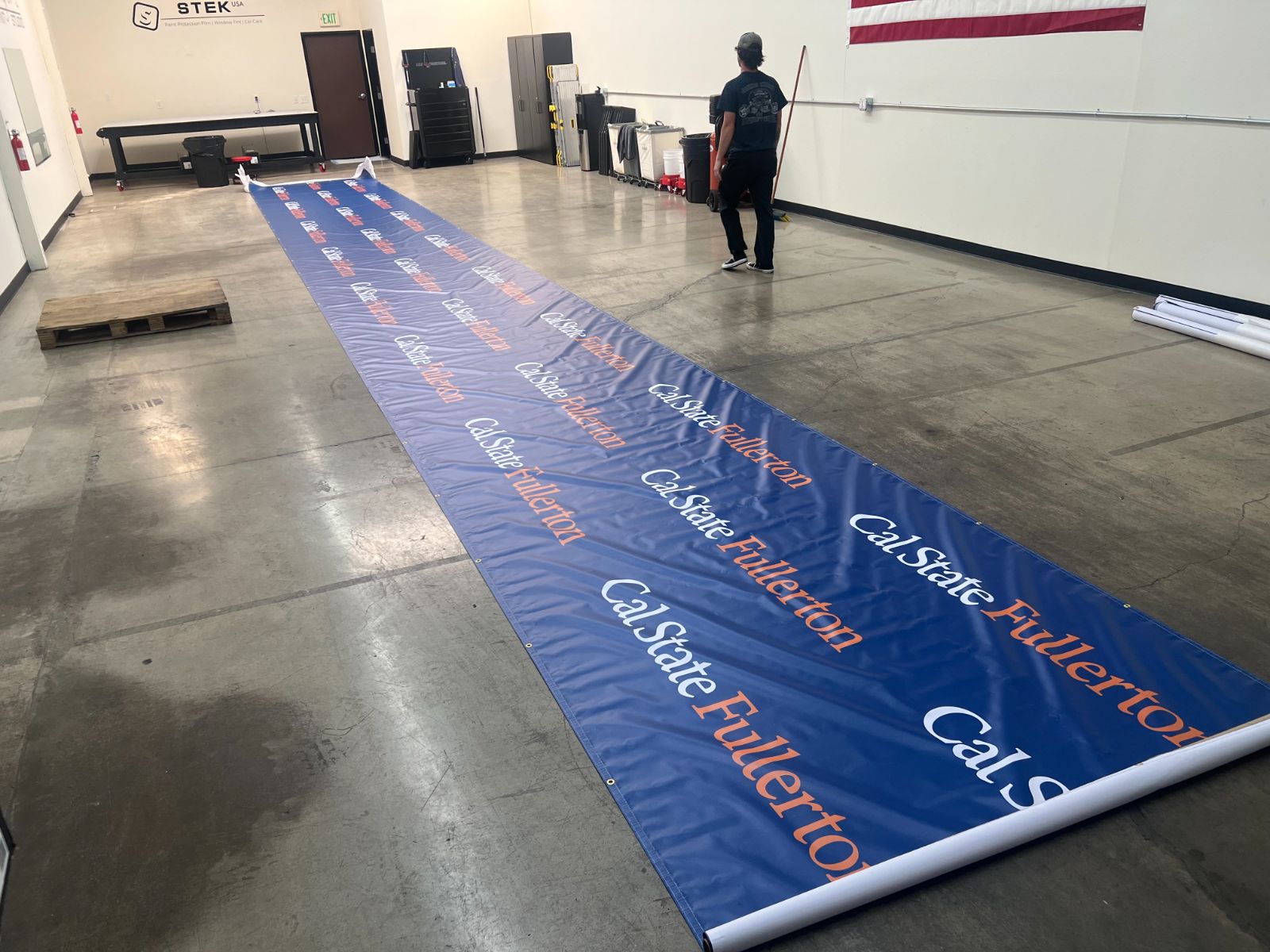 A very long Cal State Fullerton step-and-repeat banner unrolled across a shop floor: blue printed vinyl with the university wordmark repeated in white and orange, grommeted along the edges, made by Hi Sign, Brea, CA