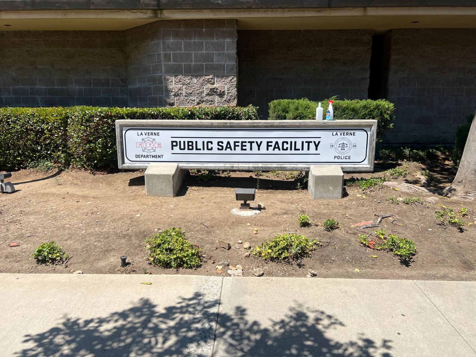 Low monument sign reading 'Public Safety Facility' set into a concrete base outside a masonry building: black text with a La Verne Fire Department crest on the left and a La Verne Police seal on the right, installed by Hi Sign, Brea, CA