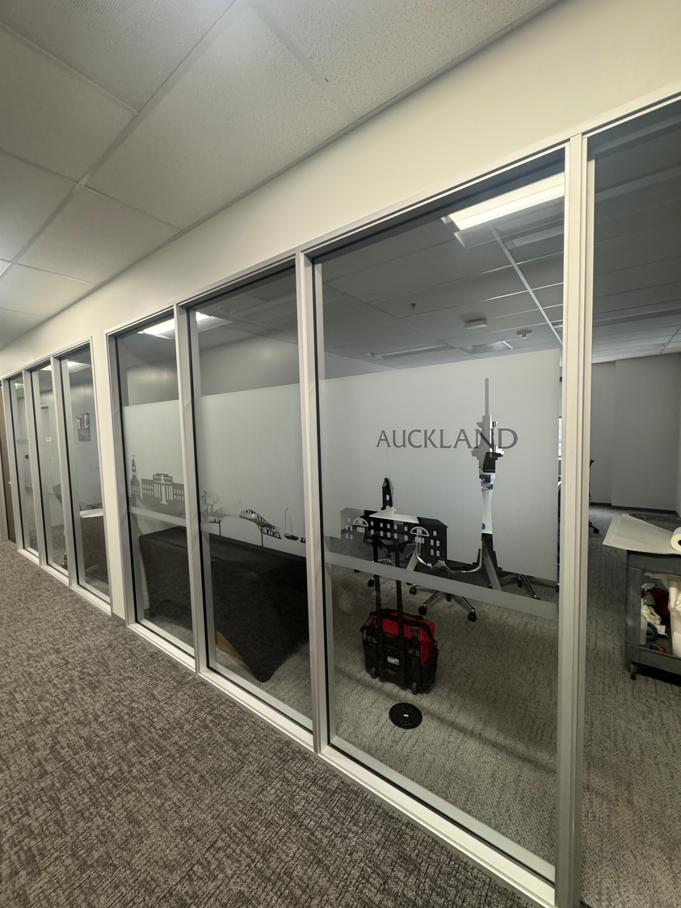 Corridor of glass-walled meeting rooms, each with a frosted privacy band carrying a cut-vinyl city skyline and room name, including AUCKLAND with its harbour bridge and tower, installed by Hi Sign, Brea, CA