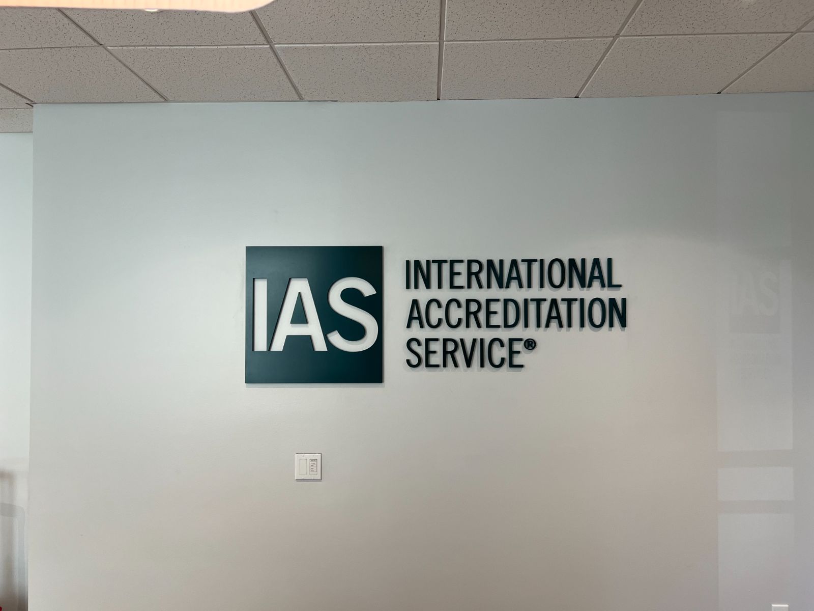 IAS (International Accreditation Service) dimensional lobby logo: dark teal ACM backer panel with raised white cast acrylic letterforms, flanked by dimensional near-black painted letters spelling 'International Accreditation Service,' standoff-mounted on a white interior office wall, installed by Hi Sign, Brea, CA