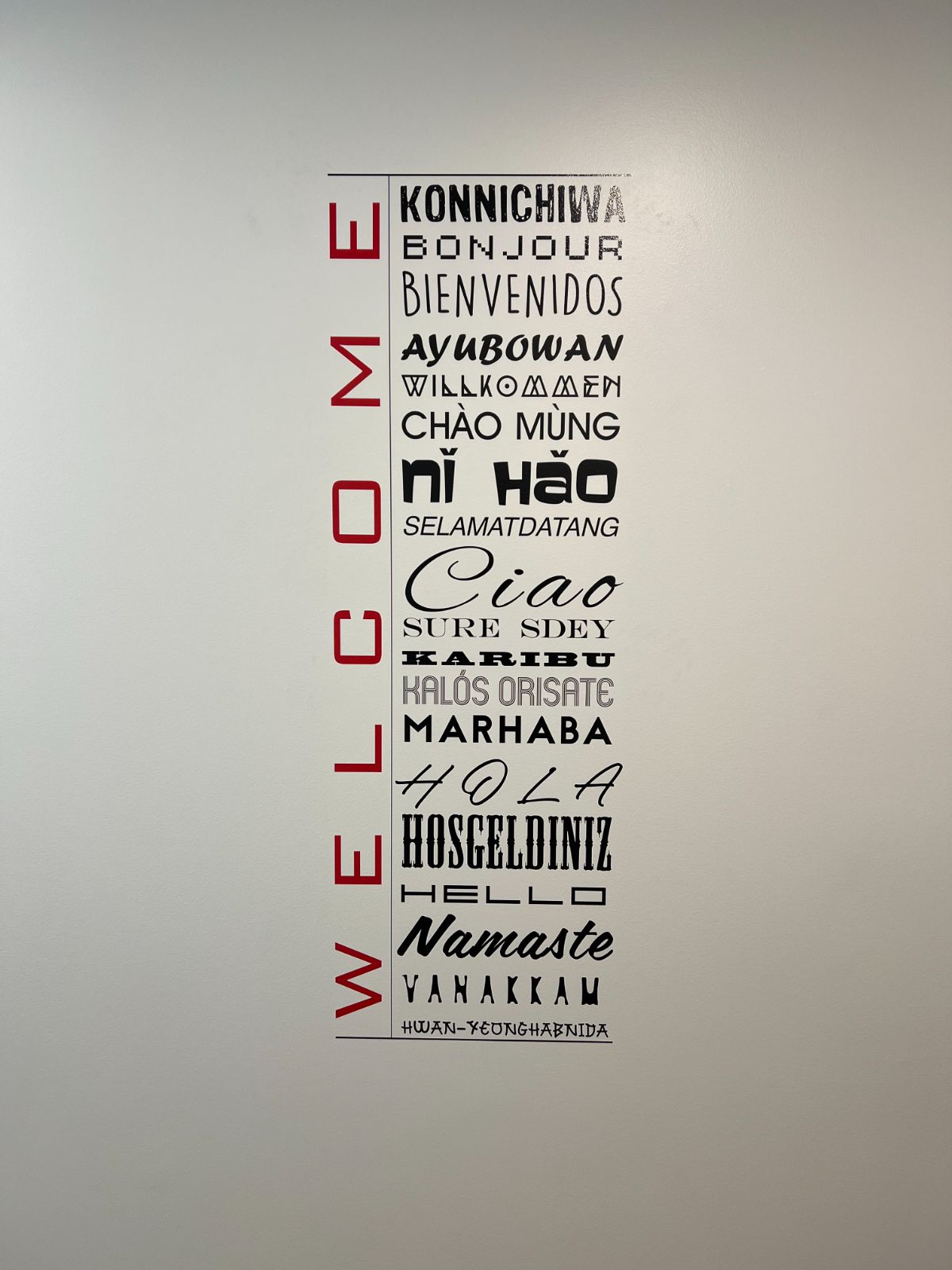 Multi-language welcome wall graphic: vertical red cut vinyl 'WELCOME' letters beside a cut vinyl panel listing greetings in 18 languages including Konnichiwa, Bonjour, Bienvenidos, Nǐ hǎo, and Namaste, applied to a white interior wall, installed by Hi Sign, Brea, CA