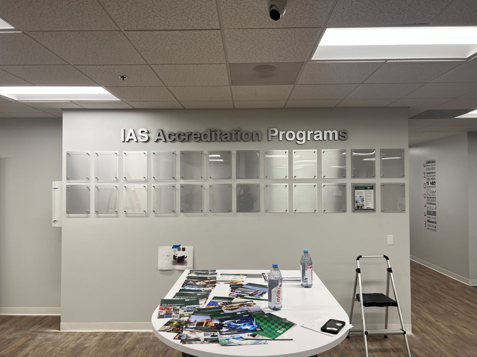 IAS Accreditation Programs corporate feature wall: large brushed aluminum dimensional letters as headline above a 4×5 grid of clear acrylic standoff-mounted document display frames on a white interior office wall, installed by Hi Sign, Brea, CA