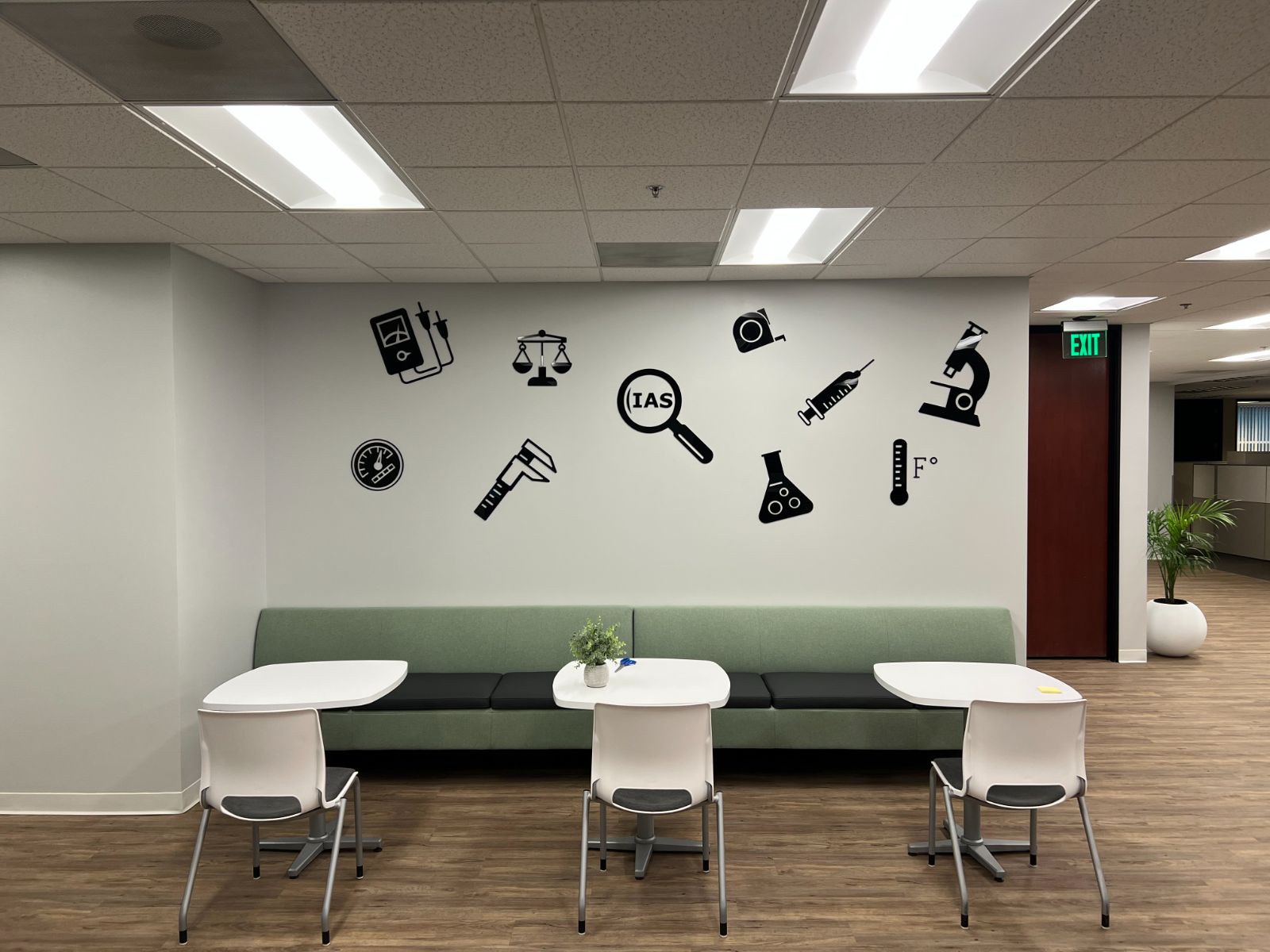 Office break area with a scatter of black cut-vinyl instrument icons on the wall: a multimeter, scale, calipers, magnifier with 'IAS', tape measure, syringe, flask, thermometer and microscope, above a green bench, installed by Hi Sign, Brea, CA