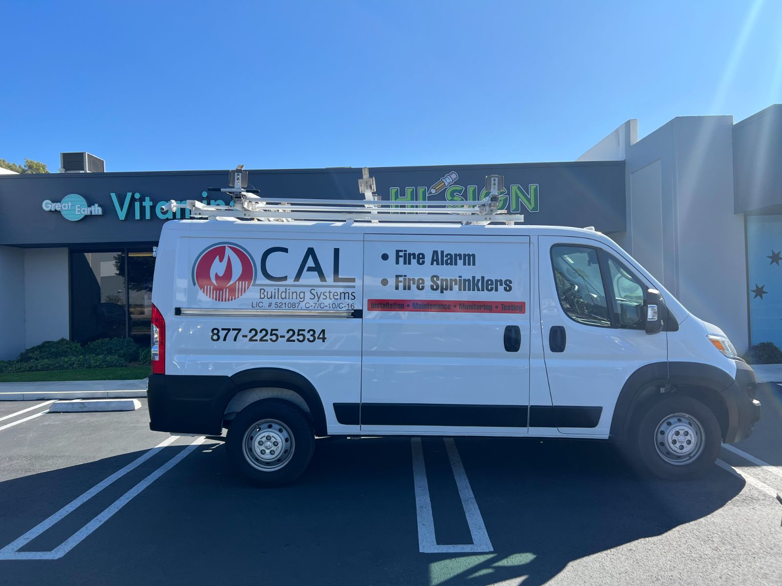 Driver side of a white CAL Building Systems cargo van parked outside the shop: cut vinyl fire-flame logo, 'Fire Alarm / Fire Sprinklers' lettering, phone number, and license numbers applied to the body panel, installed by Hi Sign, Brea, CA