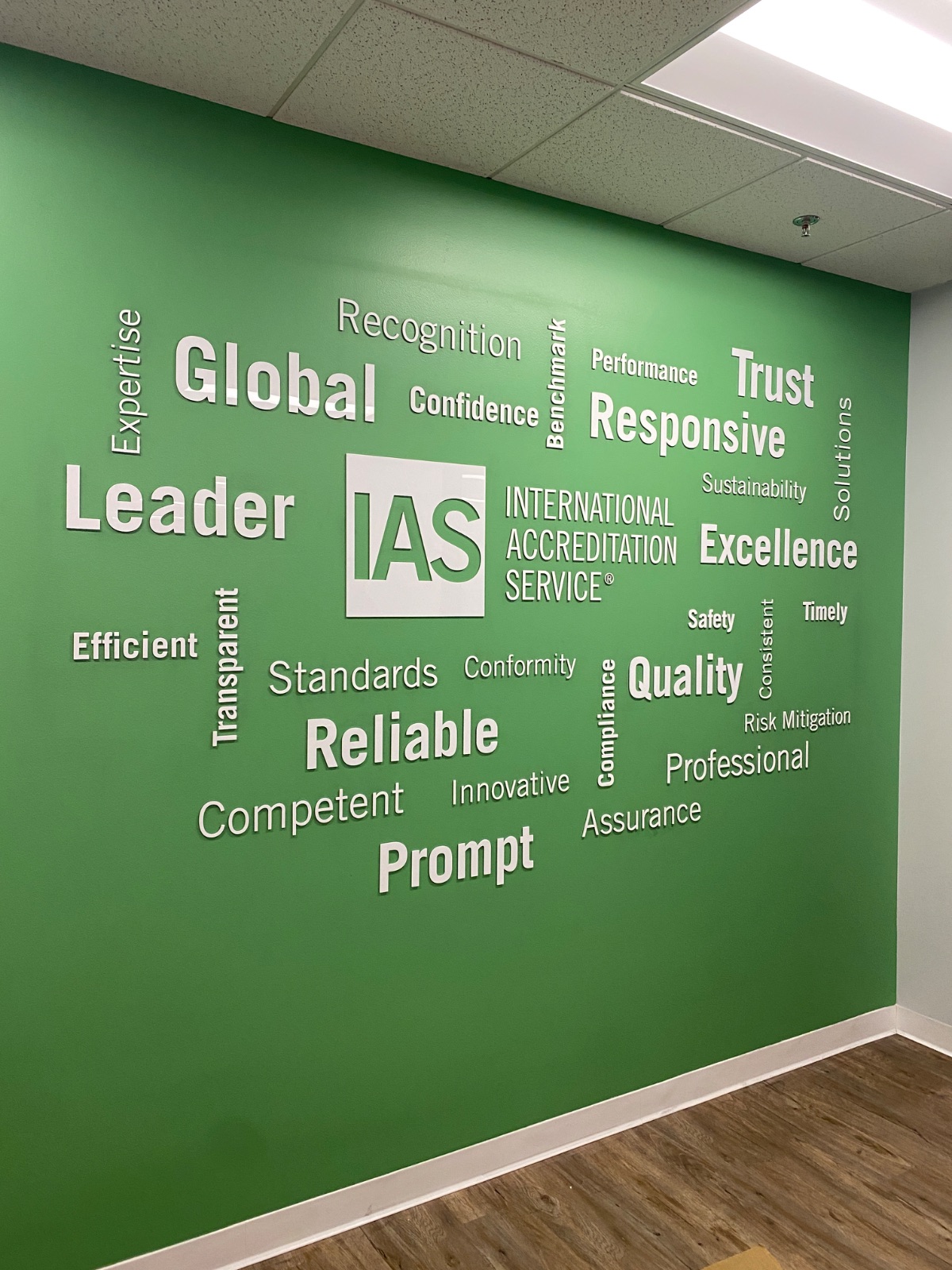 Green office wall covered in a cut-vinyl word cloud of white and grey terms (Global, Leader, Trust, Responsive, Excellence, Reliable, Prompt) around the IAS logo, installed by Hi Sign, Brea, CA