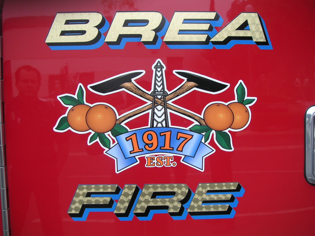 Door of a red fire engine with 'BREA' and 'FIRE' in gold-leaf-look dimensional lettering above and below a printed Brea Fire crest of crossed axes, oranges, and an oil derrick with an EST. 1917 banner, installed by Hi Sign, Brea, CA