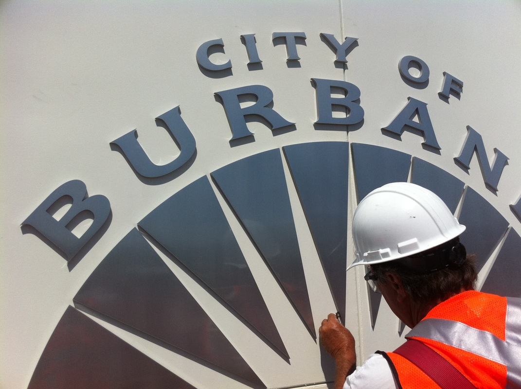 Installer in a hard hat and orange safety vest aligning gray dimensional letters spelling 'City of Burbank' above a large sunburst graphic on a white exterior panel, installed by Hi Sign, Brea, CA