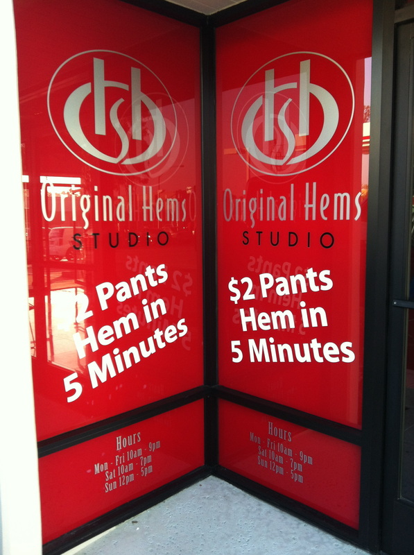 Storefront entry with red vinyl window graphics for Original Hems Studio: a silver monogram, business name, white offer copy reading '$2 Pants Hem in 5 Minutes,' and store hours across two glass panels, installed by Hi Sign, Brea, CA