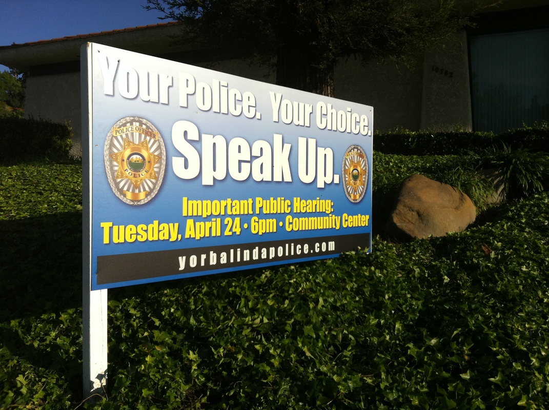 Post-mounted exterior sign on a landscaped slope reading 'Your Police. Your Choice. Speak Up.' with two police badges and public hearing details, for Yorba Linda Police, installed by Hi Sign, Brea, CA